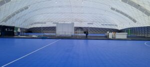 Indoor sports hall with a bright blue court, white quilted roof, and a person near equipment at center.