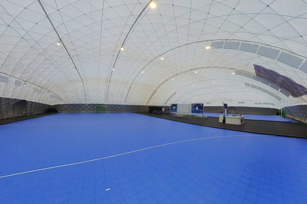 Empty indoor sports arena with a bright blue court inside a large white domed structure.