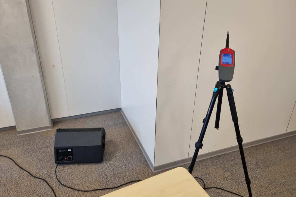 Tripod with a red handheld electronic device mounted in a corner office space; cables run across the carpet and a black speaker sits on the floor nearby.