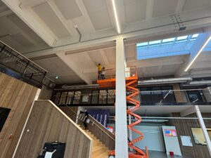 Two construction workers on an orange scissor lift reach up toward a concrete ceiling in a large industrial space with skylights and exposed pipes above them.