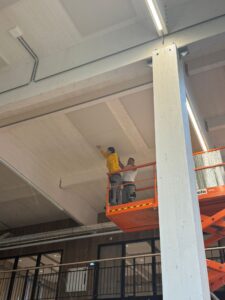 Three construction workers on an orange lift inspect and work on a high ceiling inside a building with exposed wooden beams and ducts nearby.