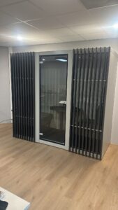 Glass door set in a small enclosed booth with vertical black slats on both sides, in a modern office with wood flooring; a table and chairs visible through the glass.